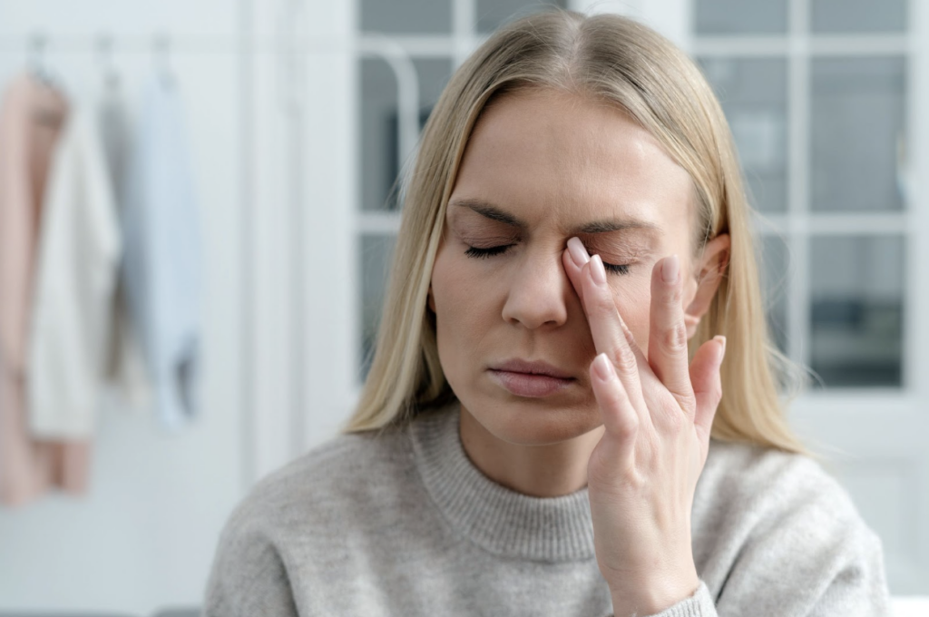 A woman with closed eyes gently touches and winces in pain at her dry eyes